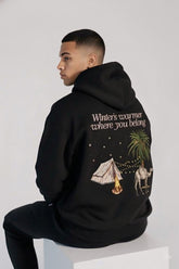 Person wearing a black hoodie with a graphic design and text on the back, sitting on a white surface.